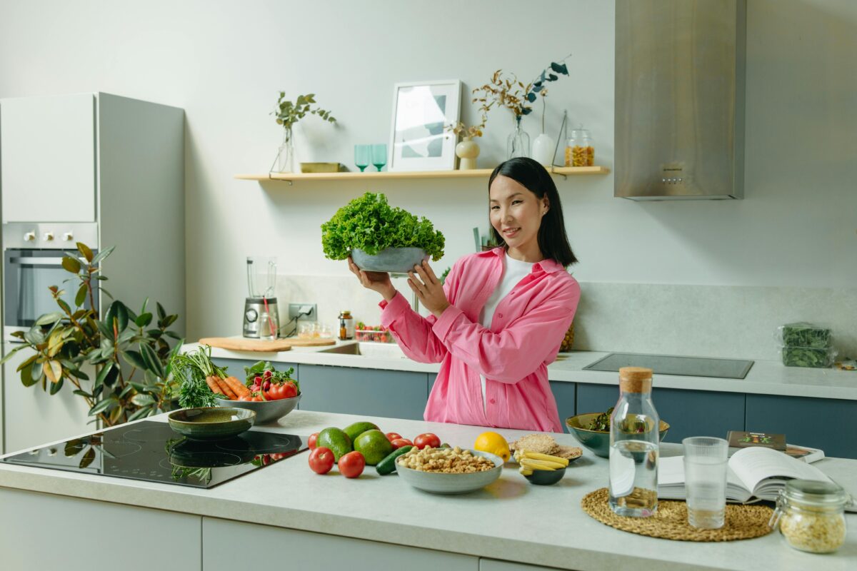 Woman in a kitchen holding fresh lettuce, surrounded by healthy ingredients like tomatoes and avocados, promoting healthy eating.