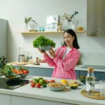Woman in a kitchen holding fresh lettuce, surrounded by healthy ingredients like tomatoes and avocados, promoting healthy eating.