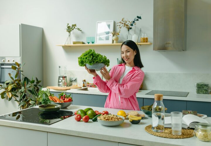 Woman in a kitchen holding fresh lettuce, surrounded by healthy ingredients like tomatoes and avocados, promoting healthy eating.